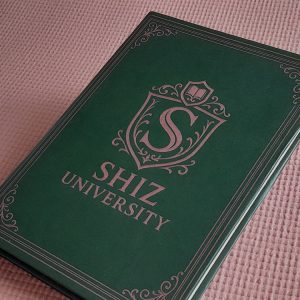 Caderno verde-escuro com capa dura e detalhes ornamentais em rosa metálico, exibindo o brasão da “Shiz University” ao centro. O fundo é uma superfície rosa texturizada.