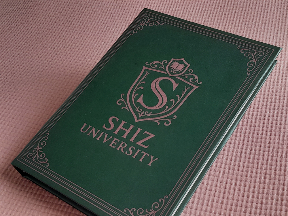 Cadernos WICKED Caderno verde-escuro com capa dura e detalhes ornamentais em rosa metálico, exibindo o brasão da “Shiz University” ao centro. O fundo é uma superfície rosa texturizada.