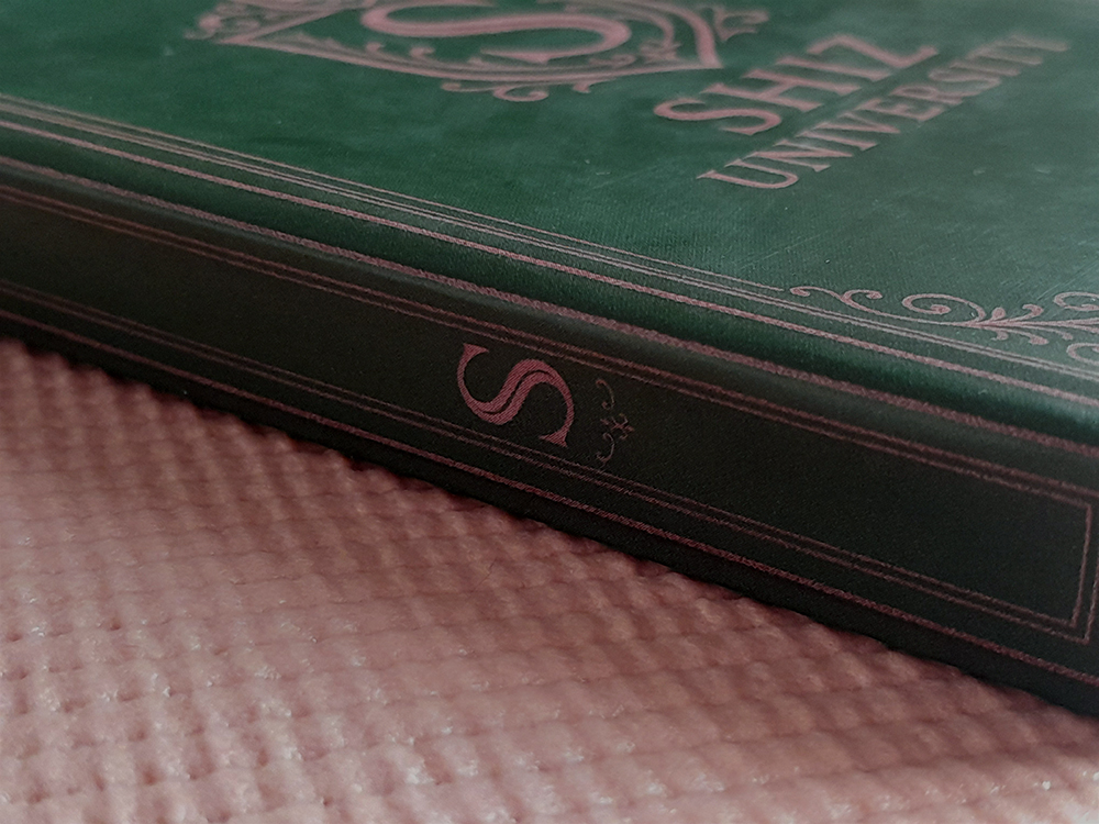 Cadernos WICKED Detalhe da lombada do caderno, onde há a letra “S” ornamentada em rosa metálico. O fundo continua sendo a base rosa texturizada.