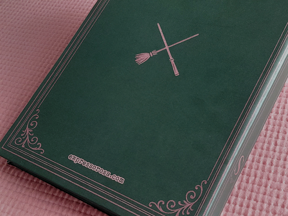 Cadernos WICKED Parte traseira do caderno, com o símbolo de uma vassoura e uma varinha cruzadas no centro, o endereço “expressorosa.com” na parte inferior e os mesmos arabescos decorativos da frente.