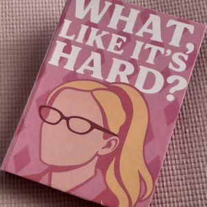 Foto de um caderno de capa dura em tons de rosa, apoiado sobre uma superfície texturizada também rosa. A capa traz a frase WHAT, LIKE IT’S HARD? em letras grandes brancas, ocupando a parte superior. Abaixo, há uma ilustração minimalista de uma mulher loira de óculos sem traços faciais, representada em estilo vetorial, remetendo à personagem Elle Woods. O fundo da capa tem um padrão geométrico em diferentes tonalidades de rosa, criando contraste suave e estético. O caderno está levemente inclinado na diagonal, destacando sua capa vibrante e temática.